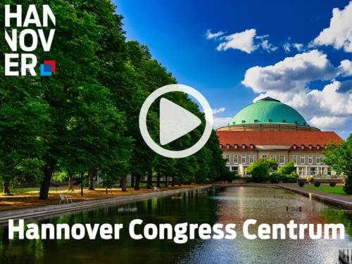 Hannover Congress Centrum mit gr&uuml;ner Kuppel, rotem Dach und spiegelndem Wasserbecken, umgeben von B&auml;umen unter blauem Himmel.